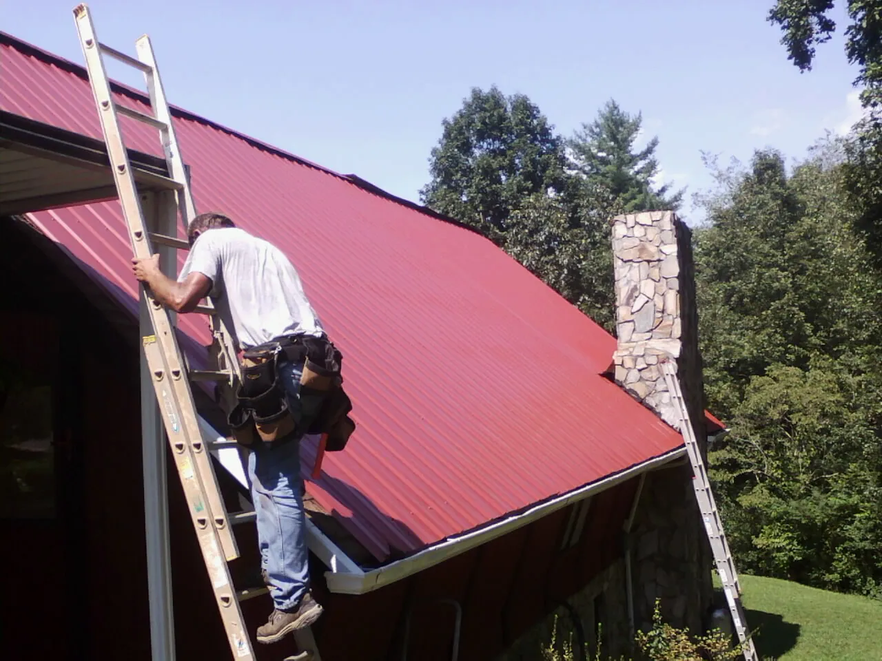 Roofer on a ladder beside a red metal roof and stone chimney — Soffit & Fascia Repair by Roofing Brothers in Scottsmoor